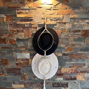 May include: A white hat with colorful paint splatters hangs on a wall with a black hat above it. Both hats are hanging from a macrame rope with a tassel.
