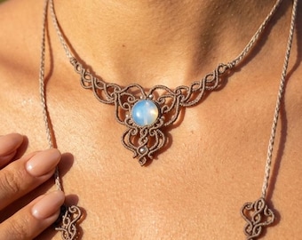 May include: A macrame necklace with a central, round, opalescent stone. The necklace features intricate knotting in a neutral color, with adjustable cords on each side. The design is centered on the chest.