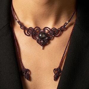 Pode incluir: Um colar gargantilha de macramé em bordeaux escuro com um padrão intrincado de cordão entrançado e uma pedra preciosa redonda e escura ao centro. O design inclui dois cordões longos e pendentes com pontas decorativas a condizer.