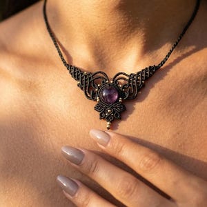 May include: A black macrame necklace featuring a central round purple amethyst gemstone. The intricate knotted cord design extends outwards from the stone, accented with small metallic beads integrated into the woven pattern around the centre.