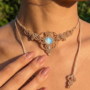Könnte beinhalten: Beige Makramee-Halskette mit einem zentralen Anhänger mit einem runden, opalisierenden Stein. Die Halskette hat aufwendige Webdetails und eine zarte, verstellbare Kordel. Das Design ist symmetrisch und elegant, passend für verschiedene Anlässe.