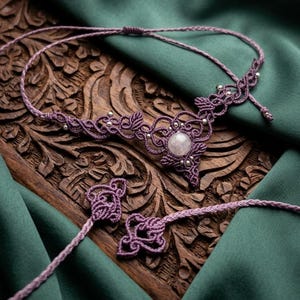 May include: A purple macramé necklace featuring a central round rose quartz gemstone, intricate knotted cord patterns, and small silver-toned beads. The necklace includes adjustable braided cords with decorative knotted ends for a secure fit.