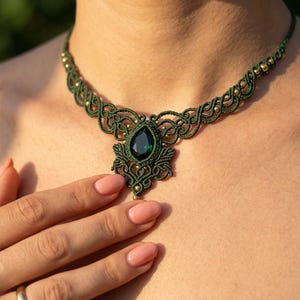 Puede incluir: Un collar de macramé verde hecho a mano con un colgante de piedra preciosa verde oscuro en forma de lágrima. El collar presenta intrincados nudos y pequeñas cuentas doradas. Una pieza de joyería única.