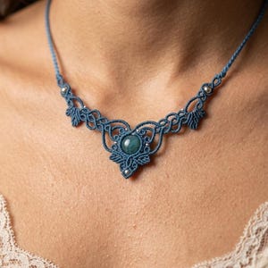 May include: A blue macrame necklace featuring a central round teal gemstone surrounded by intricate knotted cord patterns and small silver-toned beads. The design includes symmetrical leaf-like shapes and a thin cord band that encircles the neck.
