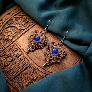 Può includere: Un paio di orecchini in macramè con intricati nodi di corda marrone a forma di goccia, ciascuno centrato con una perla blu rotonda, liscia e traslucida, completati da ganci in metallo argentato per orecchie forate.