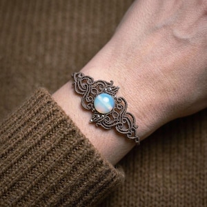 Könnte beinhalten: Ein braunes Makramee-Armband mit einem opalisierenden, runden Stein in der Mitte. Das Armband hat ein kompliziertes, offenes Muster mit kleinen silberfarbenen Perlen. Das Armband wird an einem Handgelenk gezeigt.