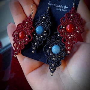 Può includere: Orecchini in macramè fatti a mano in rosso e nero. Gli orecchini rossi presentano un centro in pietra rossa, mentre gli orecchini neri hanno un centro in pietra blu. Gli orecchini hanno un design dettagliato e traforato con piccole perline d'argento. La carta dice "Handmade with love."