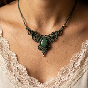 Op de afbeelding: Een groene macramé ketting met een druppelvormige groene edelsteen hanger. Het ingewikkelde knoopwerkontwerp is versierd met goudkleurige kralen. De ketting wordt gedragen door een persoon die een witte kanten top draagt.