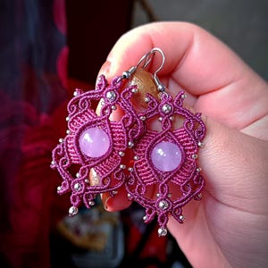 Puede incluir: Un par de pendientes de color morado y plateado con un diseño delicado e intrincado. Los pendientes presentan una piedra preciosa redonda de color morado claro en el centro de cada pendiente.