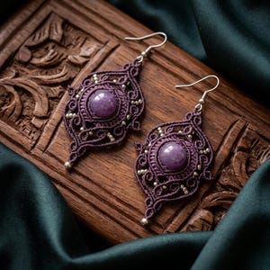 Peut inclure: Une paire de boucles d'oreilles en macramé violet foncé faites à la main, avec une pierre précieuse violette ronde au centre. Les boucles d'oreilles présentent un design ajouré détaillé avec des accents argentés et des crochets d'oreilles. Elles sont présentées sur une surface en bois.