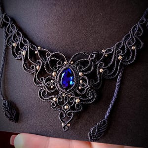 May include: A handcrafted black macrame necklace with a large, oval, deep blue gemstone center. The intricate design features delicate knotwork and small gold-colored beads, creating an elegant, bohemian style statement piece.