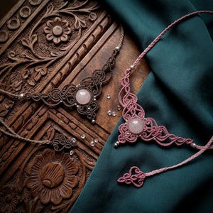 Puede incluir: Dos collares de macramé con intrincados patrones de cordón anudado y cabujones redondos de cuarzo rosa en el centro. Un collar está hecho de cordón marrón oscuro y el otro de cordón rosa empolvado, ambos con pequeñas cuentas plateadas.