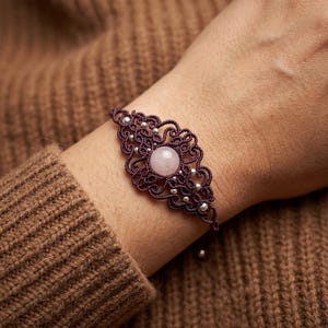 May include: A dark purple macrame bracelet with a central rose quartz stone and small silver beads. The bracelet is intricately woven with a decorative pattern. The bracelet is shown on a wrist.