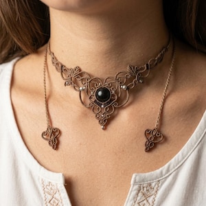 May include: A tan macrame choker necklace featuring an intricate knotted design with a central round black bead and small silver-toned accents. The necklace includes two long braided cords hanging down, each finished with a decorative knotted end.