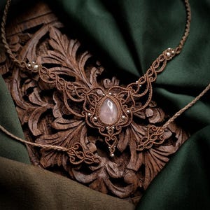 May include: A handmade macramé necklace featuring a rose quartz gemstone at its centre. The brown knotwork is detailed with small silver beads. The necklace is displayed on a carved wooden surface with a green fabric backdrop.