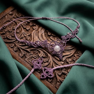 Könnte beinhalten: Eine violette Makramee-Halskette mit einem zentralen runden Rosenquarz, komplizierten geknoteten Kordelmustern und kleinen silberfarbenen Perlen. Die Kette verfügt über dünne geflochtene Kordeln mit dekorativen Knotenenden.