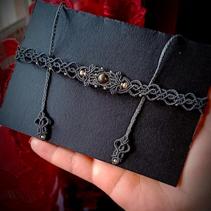 Op de afbeelding: Een handgemaakte macramé choker ketting in donkergrijs, met ingewikkeld knoopwerk en kleine goudkleurige kralen. De ketting heeft een centraal ontwerp met een grotere kraal en delicate bungelende accenten. De choker wordt gepresenteerd op een zwart oppervlak.