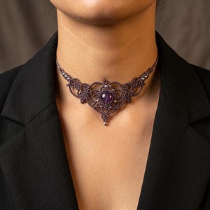 Może przedstawiać: Ciemnobrązowy naszyjnik typu choker wykonany techniką makramy, z misternym wzorem wiązanych sznurków, centralnie umieszczonym okrągłym fioletowym kamieniem szlachetnym oraz małymi metalicznymi srebrnymi koralikami wplecionymi w symetryczny wzór.