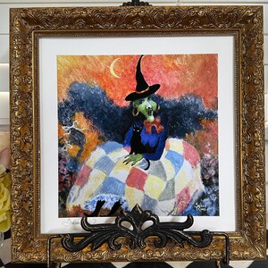 May include: A square painting in an ornate gold frame featuring a green-skinned witch wearing a black hat and blue top, sitting with a black cat on a colorful patchwork quilt against an orange sky with a crescent moon, displayed on a black stand.