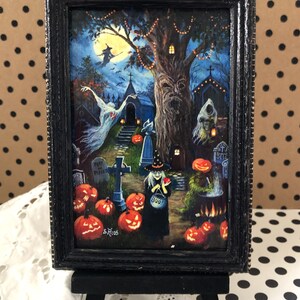May include: A framed Halloween-themed painting depicting a witch with a cauldron, a ghost, and a spooky tree with jack-o'-lanterns. The painting is set against a dark blue background with a full moon and stars. The text "Rest in Pieces" and "Boo!" are visible on the painting.
