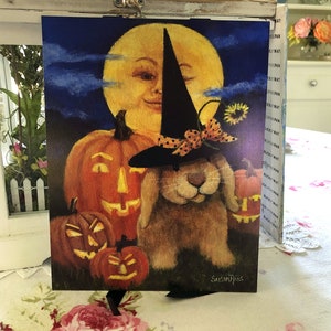 May include: A Halloween-themed painting featuring a bunny wearing a witch's hat and holding a pumpkin. The bunny is surrounded by pumpkins and a smiling moon in the background.