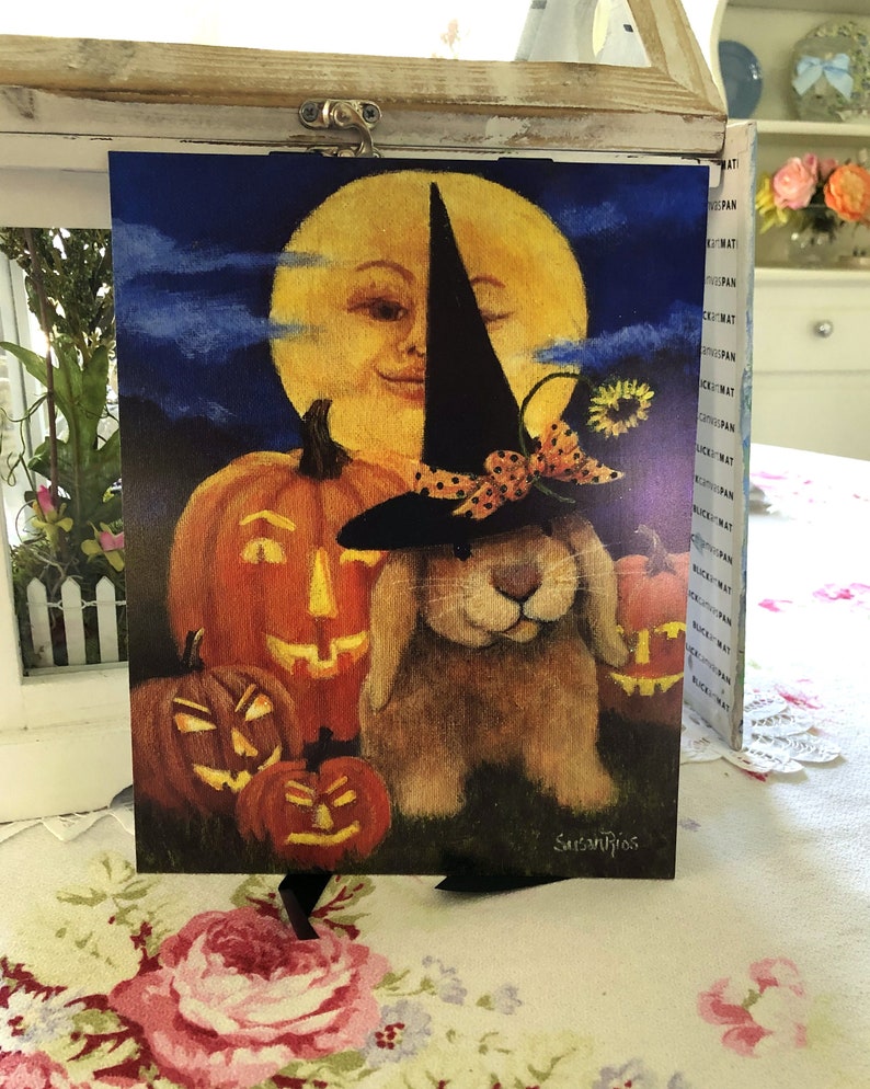 May include: A Halloween-themed painting featuring a bunny wearing a witch's hat and holding a pumpkin. The bunny is surrounded by pumpkins and a smiling moon in the background.