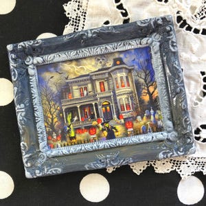 May include: A framed Halloween-themed picture. The artwork depicts a spooky haunted house with jack-o'-lanterns, witches, and bats. The frame is a distressed gray with ornate detailing. The picture is set against a black and white polka dot background.