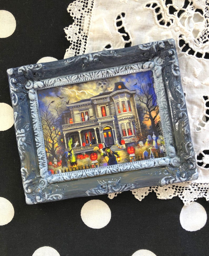 May include: A framed Halloween-themed picture. The artwork depicts a spooky haunted house with jack-o'-lanterns, witches, and bats. The frame is a distressed gray with ornate detailing. The picture is set against a black and white polka dot background.