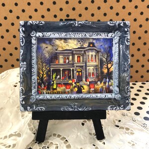 May include: A framed Halloween-themed miniature artwork. The artwork depicts a spooky haunted house scene with a graveyard, pumpkins, and figures in costumes. The frame is ornate, with a distressed gray and white finish, and is displayed on a small black easel.