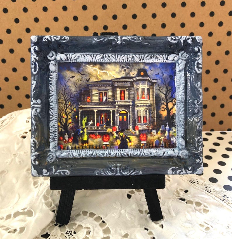 May include: A framed Halloween-themed miniature artwork. The artwork depicts a spooky haunted house scene with a graveyard, pumpkins, and figures in costumes. The frame is ornate, with a distressed gray and white finish, and is displayed on a small black easel.