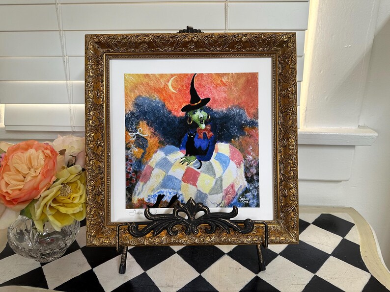 May include: A square painting in an ornate gold frame featuring a green-skinned witch wearing a black hat and blue top, sitting with a black cat on a colorful patchwork quilt against an orange sky with a crescent moon, displayed on a black stand.