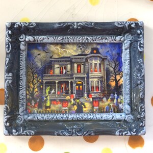 May include: A framed Halloween-themed picture featuring a spooky haunted house with lit windows, surrounded by trees, pumpkins, and tombstones. The frame is dark gray with ornate detailing, adding a vintage touch to the artwork.