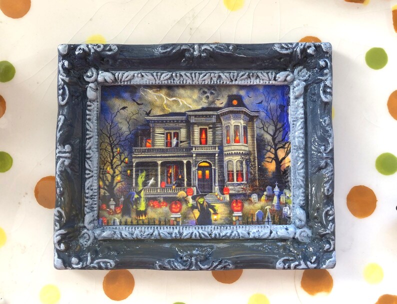 May include: A framed Halloween-themed picture featuring a spooky haunted house with lit windows, surrounded by trees, pumpkins, and tombstones. The frame is dark gray with ornate detailing, adding a vintage touch to the artwork.