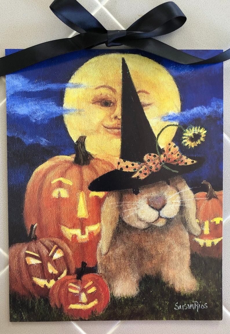 May include: A painting of a brown bunny wearing a black witch hat with a red and black polka dot ribbon. The bunny is surrounded by four orange pumpkins with carved jack-o-lantern faces. The background is a blue night sky with a yellow moon with a smiling face. The artist's signature 'Susan Rios' is in the bottom right corner.