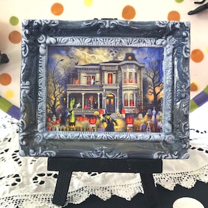 May include: A framed Halloween-themed picture of a haunted house, set on a small black easel. The picture features a spooky mansion with jack-o'-lanterns, ghosts, and a graveyard. The frame is dark gray with ornate detailing.