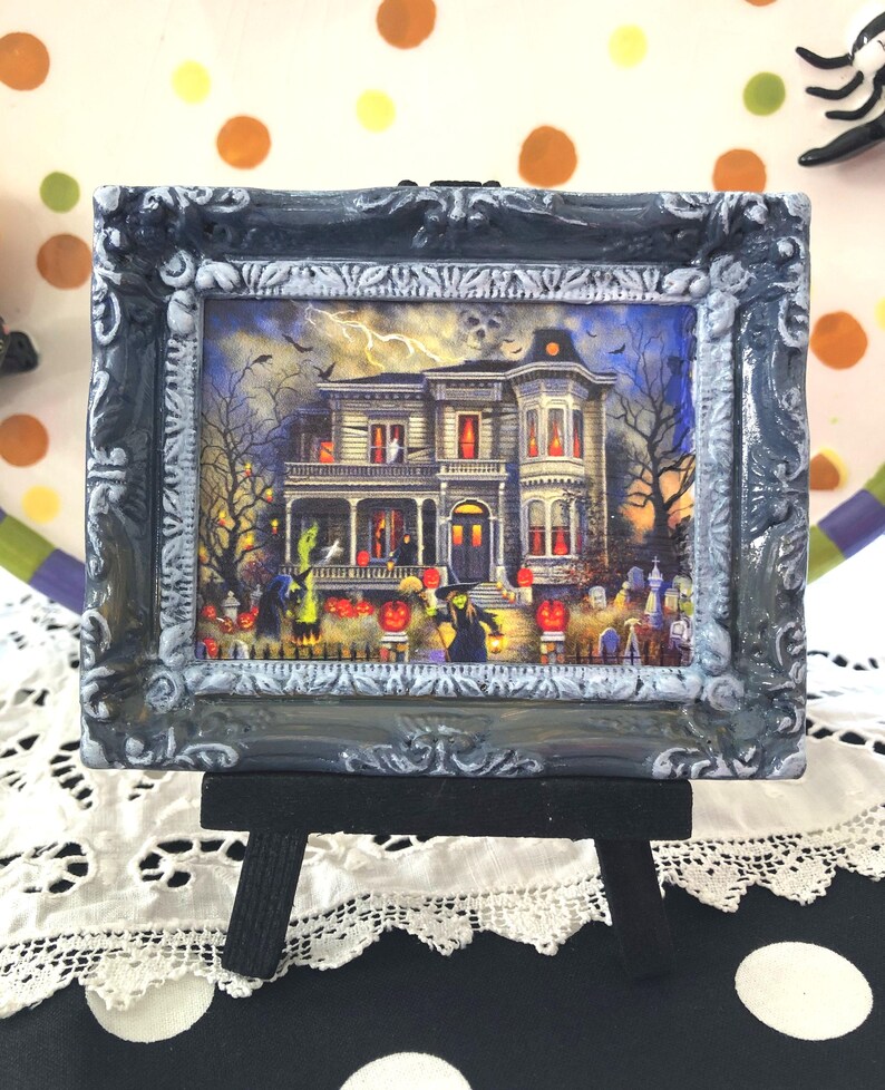 May include: A framed Halloween-themed picture of a haunted house, set on a small black easel. The picture features a spooky mansion with jack-o'-lanterns, ghosts, and a graveyard. The frame is dark gray with ornate detailing.