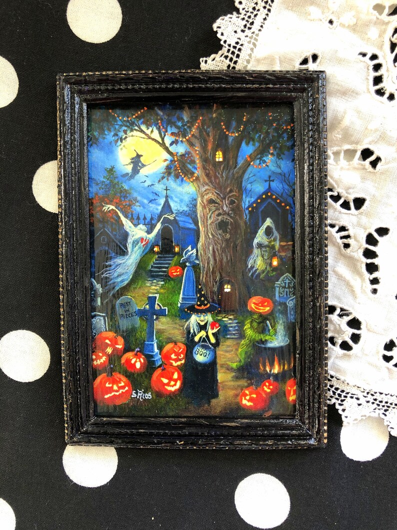 May include: A framed painting of a Halloween scene with a witch, a graveyard, and a spooky tree. The witch is holding a bucket of candy and a red apple. The graveyard has a tombstone that reads "Rest in Pieces". The tree has a face carved into it and is decorated with lights. The painting is set against a dark blue background with a full moon.