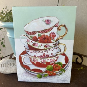 May include: A stack of four vintage teacups with floral patterns in shades of red, pink, and white. The cups are stacked on a red and white saucer with a green vine and red flowers.