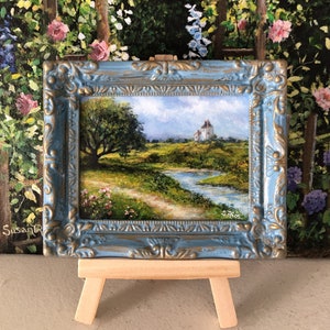 May include: A small framed painting of a landscape with a white house in the distance, a winding path, and a river. The frame is blue with gold accents and is on a small wooden easel.