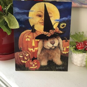 May include: A Halloween card featuring a brown bunny wearing a witch's hat and a bow. The bunny is surrounded by jack-o'-lanterns and a smiling moon face.
