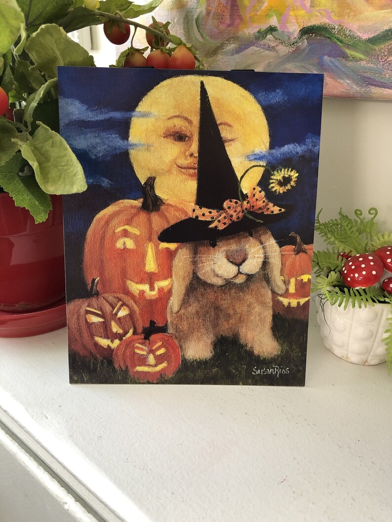 May include: A Halloween card featuring a brown bunny wearing a witch's hat and a bow. The bunny is surrounded by jack-o'-lanterns and a smiling moon face.