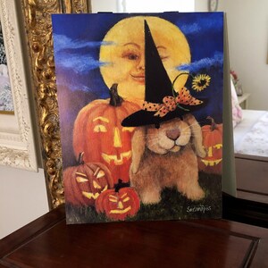 May include: A brown bunny wearing a black witch hat with a red and white polka dot bow sits in front of a full moon and three jack-o'-lanterns. The moon has a smiling face. The image is painted in a whimsical style.