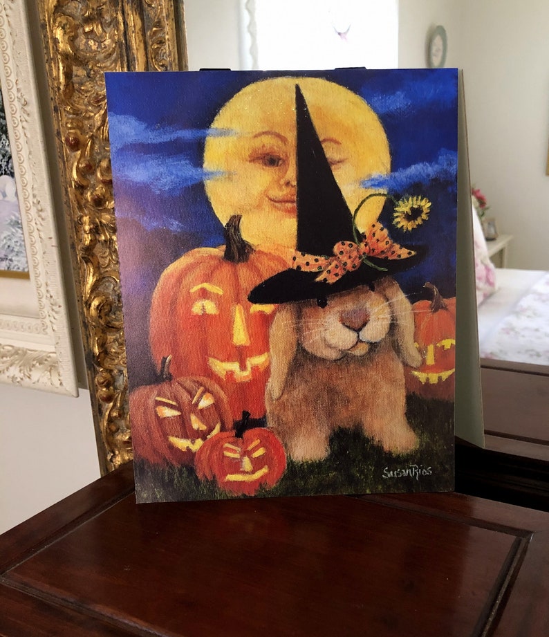 May include: A brown bunny wearing a black witch hat with a red and white polka dot bow sits in front of a full moon and three jack-o'-lanterns. The moon has a smiling face. The image is painted in a whimsical style.