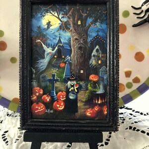 May include: A black framed painting of a Halloween scene with a witch, a graveyard, and a spooky tree. The witch is holding a bucket that says 'Boo!' and an apple. The painting is on a small black easel.