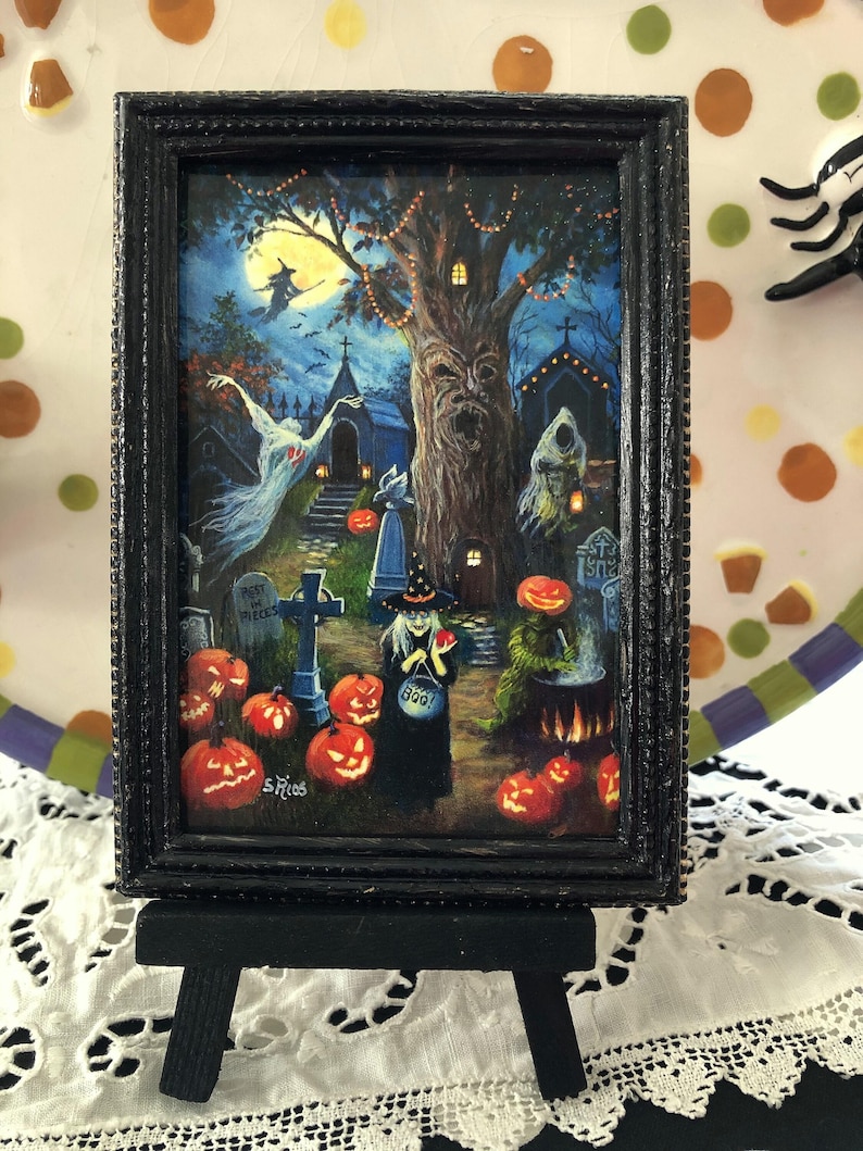 May include: A black framed painting of a Halloween scene with a witch, a graveyard, and a spooky tree. The witch is holding a bucket that says 'Boo!' and an apple. The painting is on a small black easel.