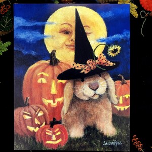 May include: A brown bunny wearing a black witch hat with orange and white polka dots and a yellow flower. The bunny is surrounded by three orange pumpkins with carved faces. The background is a dark blue sky with a yellow moon with a smiling face.