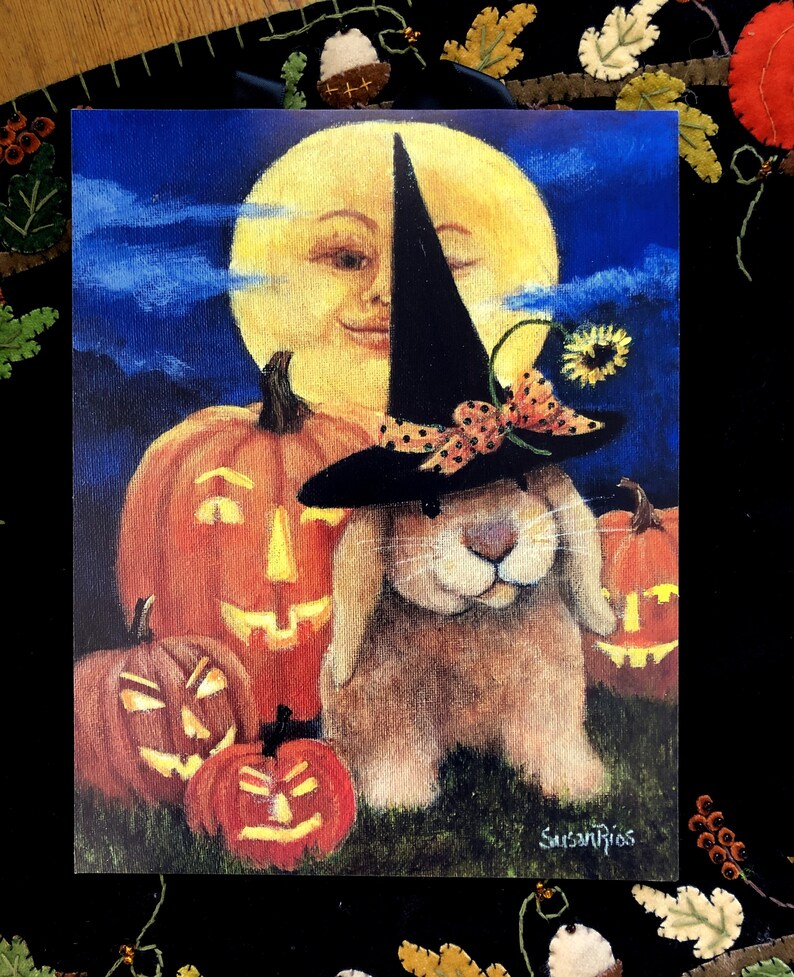 May include: A brown bunny wearing a black witch hat with orange and white polka dots and a yellow flower. The bunny is surrounded by three orange pumpkins with carved faces. The background is a dark blue sky with a yellow moon with a smiling face.