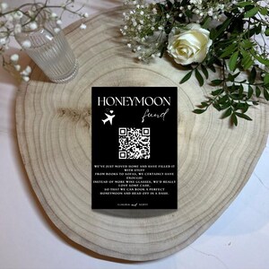 Pode incluir: Um cartão preto com as palavras "HONEYMOON fund" e um código QR. O cartão também inclui texto sobre o desejo de um casal por dinheiro em vez de presentes. O cartão está em uma superfície de madeira, com um vaso de vidro e flores ao fundo.
