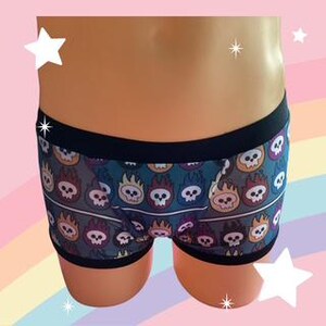 May include: Boxer briefs featuring a repeating pattern of white skulls surrounded by colorful flames in shades of orange, purple, and blue on a dark background. The garment has a solid black elastic waistband and black trim along the leg openings.
