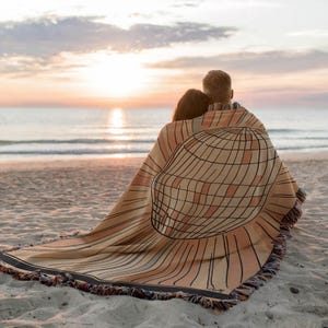 May include: A couple sits on a sandy beach, wrapped in a woven throw blanket with a geometric pattern. The blanket features a large, circular design with a grid pattern, reminiscent of a sun or a globe. The couple is facing the ocean, watching the sunset.